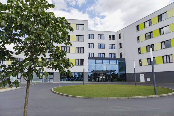 Außenansicht eines modernen Klinik- oder Pflegegebäudes mit mehreren Stockwerken, hellen Fassaden und grün akzentuierten Fenstern; davor eine begrünte Zufahrt mit Eingangsbereich.