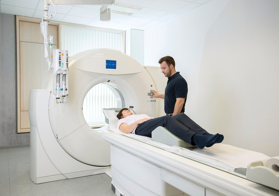 Patientin liegt auf einer Liege vor einem CT-Scanner, während ein medizinischer Mitarbeiter das Gerät vorbereitet und die Untersuchung überwacht.
