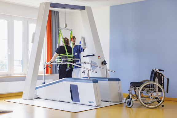 Patientin trainiert mit Unterstützung einer Therapeutin an einem Gehtrainer mit Hebevorrichtung in einem hellen Rehabilitationsraum; ein Rollstuhl steht daneben.