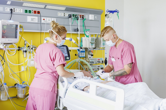 Zwei Pflegefachkräfte versorgen einen Patienten auf einer Intensivstation; sie tragen Schutzmasken und arbeiten an einem Krankenbett mit medizinischen Monitoren und Geräten.