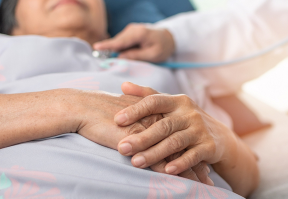 Nahaufnahme der gefalteten Hände einer Patientin, die entspannt in einem Krankenhausbett liegt; im Hintergrund misst eine Pflegekraft die Vitalwerte.