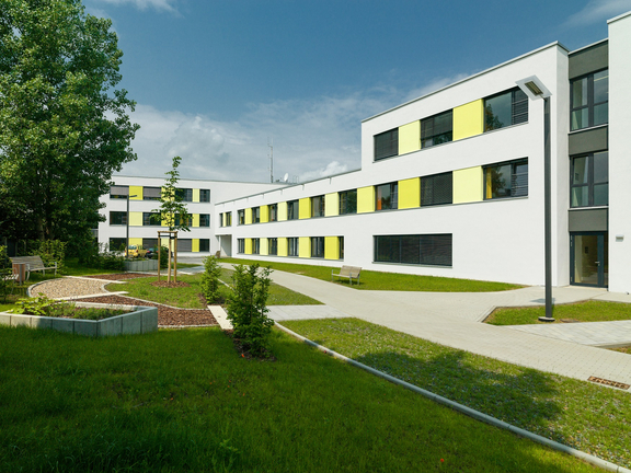 Moderner Klinik- oder Pflegegebäudekomplex mit weißer Fassade und gelb akzentuierten Fenstern, umgeben von Grünflächen, Wegen und Sitzgelegenheiten im Außenbereich.