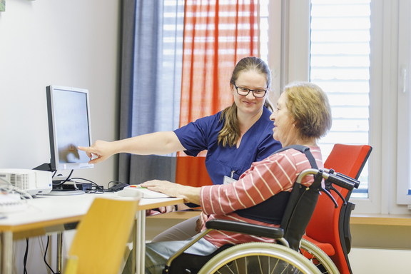 Therapeutin erklärt einer älteren Frau im Rollstuhl eine Übung am Computer; sie zeigt auf den Bildschirm in einem hellen Therapieraum.