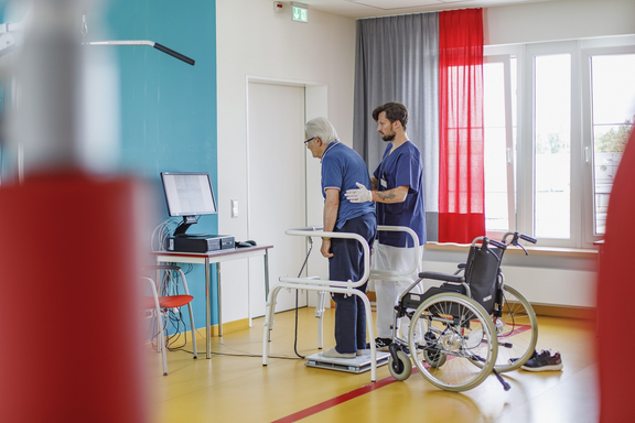 Therapeut begleitet einen älteren Mann beim Stehen und Training an einer Gehhilfe in einem Therapieraum; ein Rollstuhl steht bereit daneben.