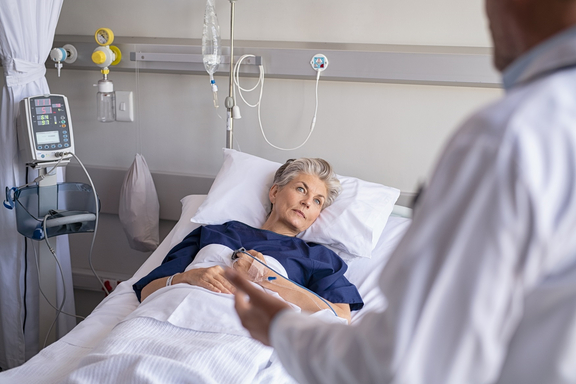 Patientin liegt in einem Krankenhausbett und blickt aufmerksam zu einem Arzt, der neben dem Bett steht; medizinische Geräte und Infusionen sind im Hintergrund zu sehen.