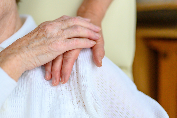 Nahaufnahme zweier Hände: Eine Pflegekraft hält beruhigend die Hand einer älteren Person auf deren Schulter.