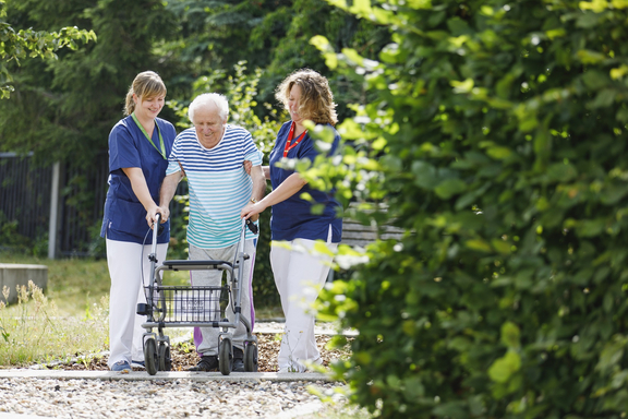Zwei Pflegekräfte begleiten einen älteren Mann beim Gehen mit einem Rollator auf einem Weg im Grünen; sie unterstützen ihn links und rechts in ruhiger Umgebung.